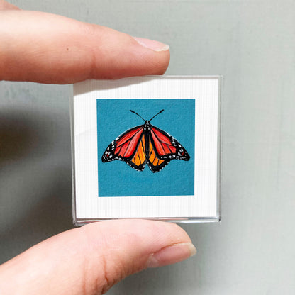 A person's hand is holding a small square with a vibrant image of a butterfly on it.