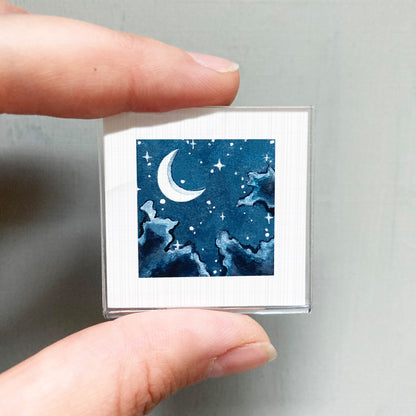 A person's hand holding a small square picture depicting a night sky with a crescent moon and stars.