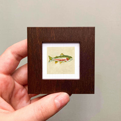 A hand is holding a small wooden frame containing a framed illustration of a fish.