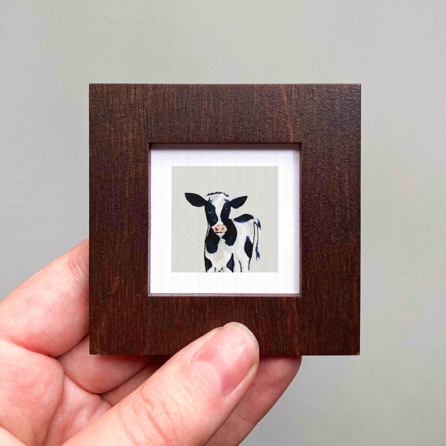 A hand is holding a small wooden frame containing a picture of a black and white cow.