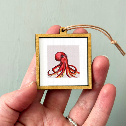 A person's hand holding a small wooden frame containing a picture of an octopus.