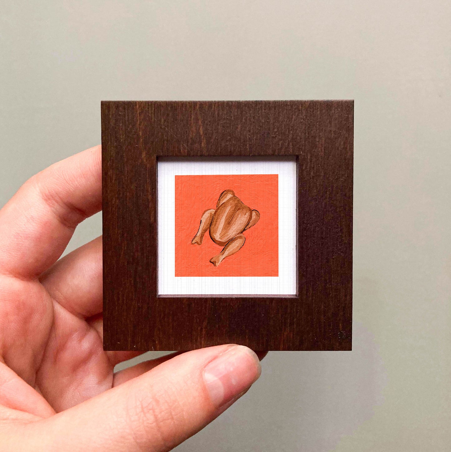A hand is holding a small wooden frame containing a framed illustration of a roasted chicken.