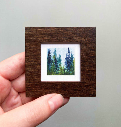 A hand is holding a small, square-shaped wooden frame containing a painting of a forest scene with tall trees.