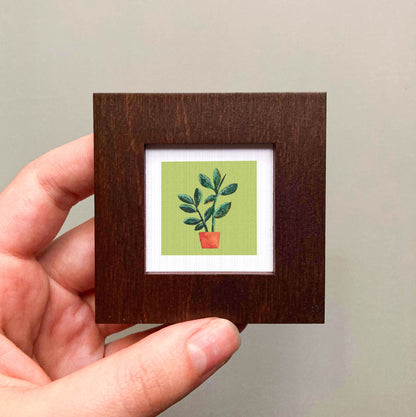 A hand is holding a small wooden frame containing a framed illustration of a potted plant with green leaves.
