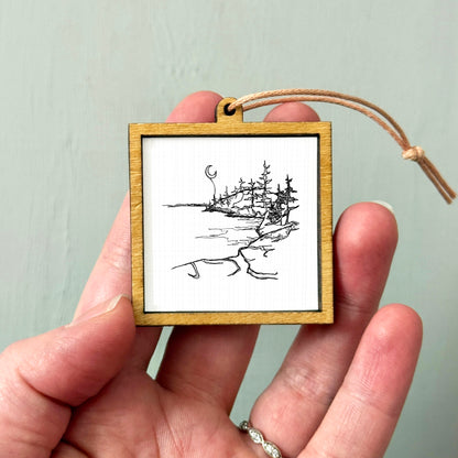 A hand holding a small wooden frame containing a black and white sketch of a landscape with trees and a crescent moon.