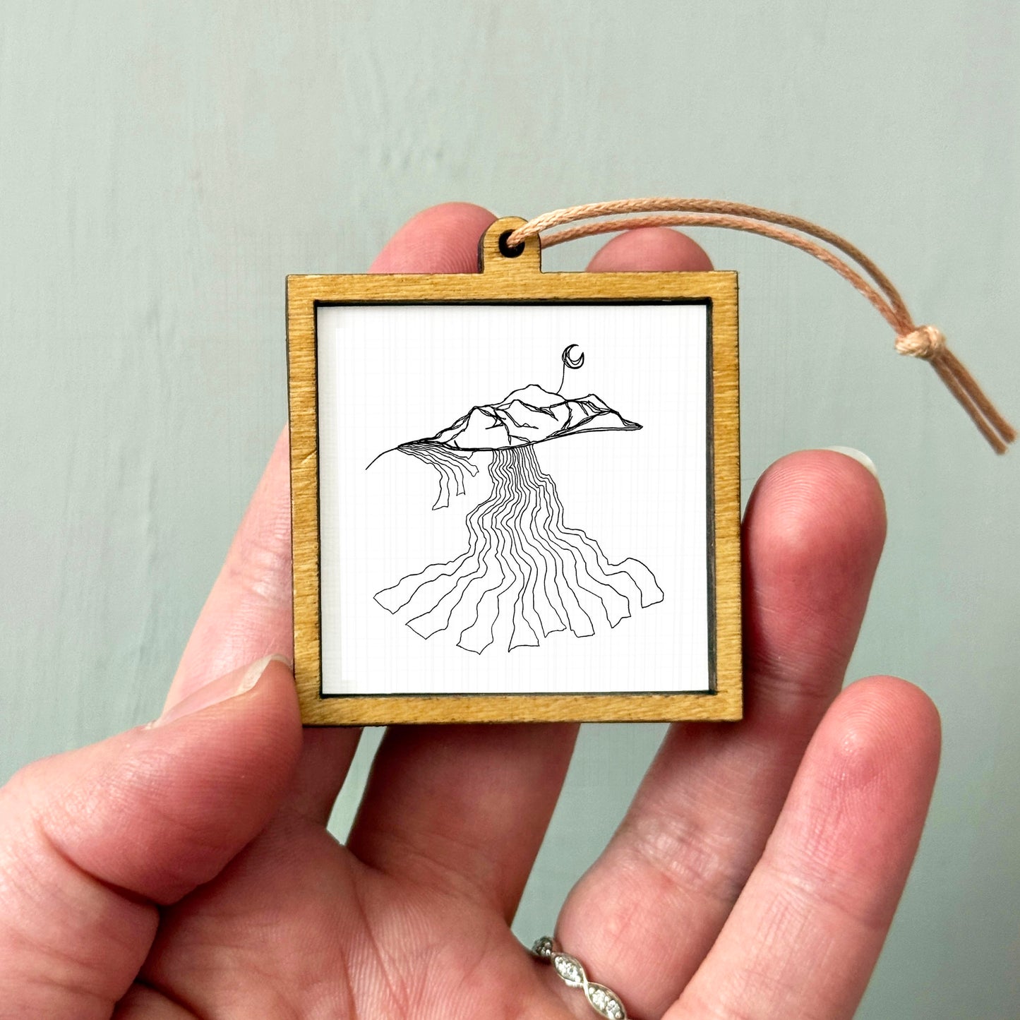 A hand holding a small wooden frame containing a black and white line drawing of a mountain landscape.