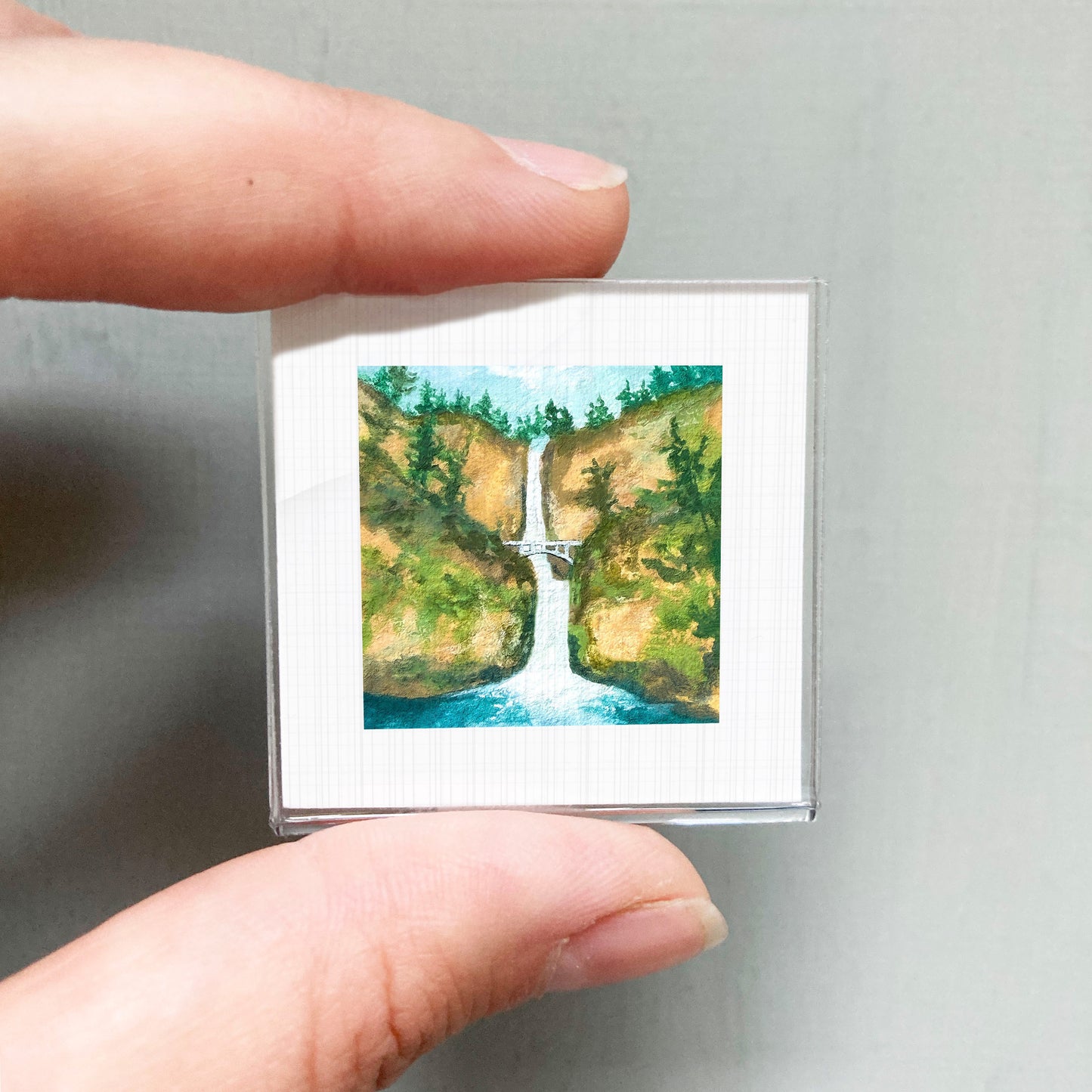 A person's hand holding a small square picture of a waterfall surrounded by lush greenery.