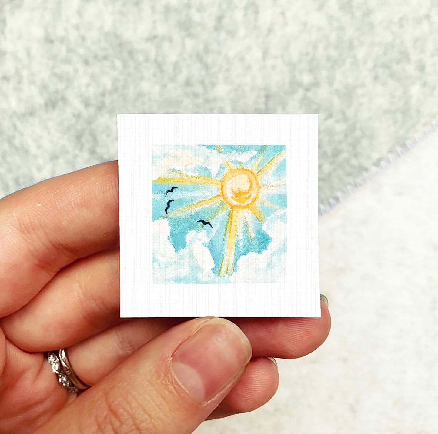 A hand holding a small square artwork depicting a sun with clouds and birds in the sky.