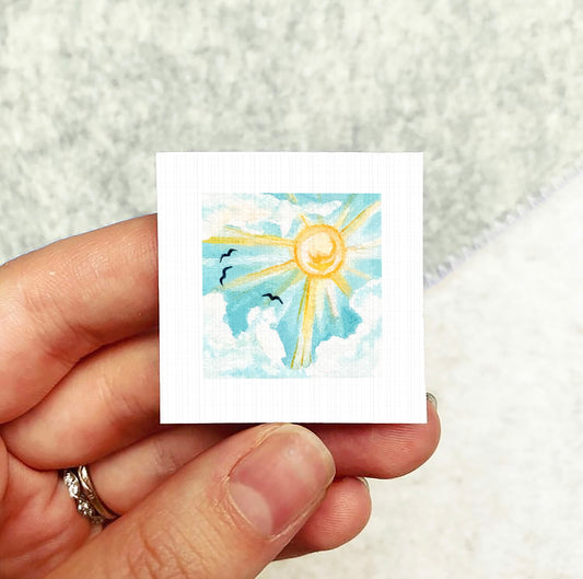 A hand holding a small square artwork depicting a sun with clouds and birds in the sky.