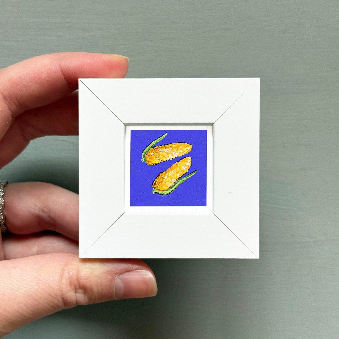 A hand is holding a small white square frame containing a vibrant painting of corn on the cob against a blue background.