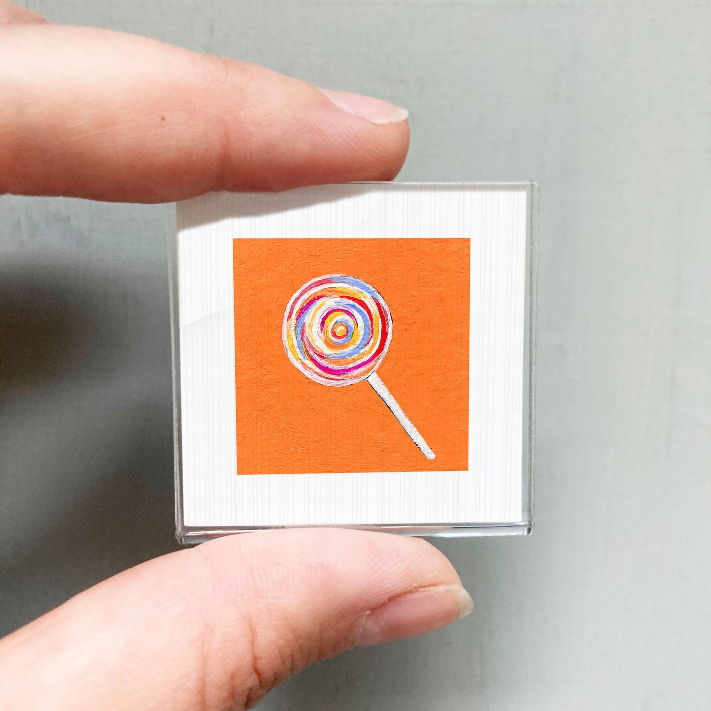 A person's hand holding a small square with an orange background and a colorful lollipop illustration on it.
