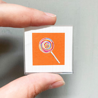 A person's hand holding a small square with an orange background and a colorful lollipop illustration on it.