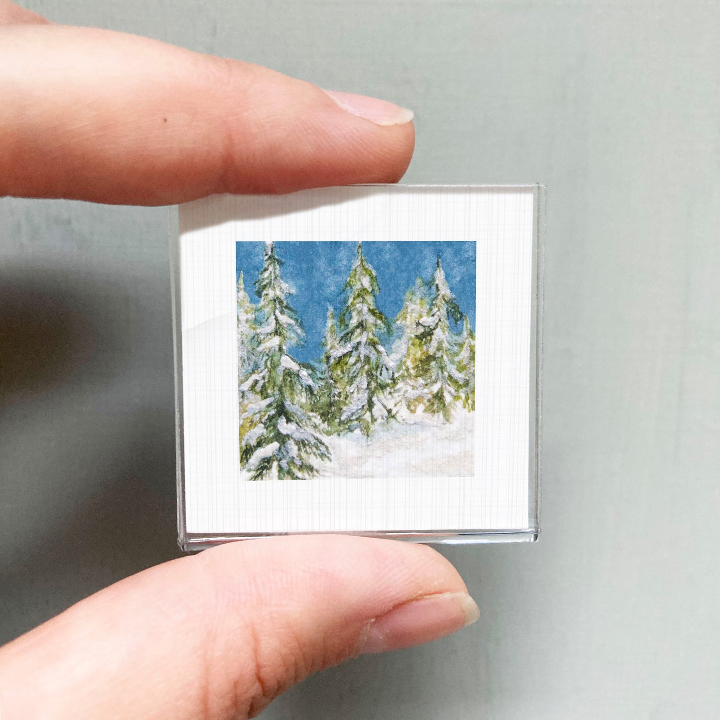 A person's hand holding a small square picture of a snowy winter landscape with pine trees.