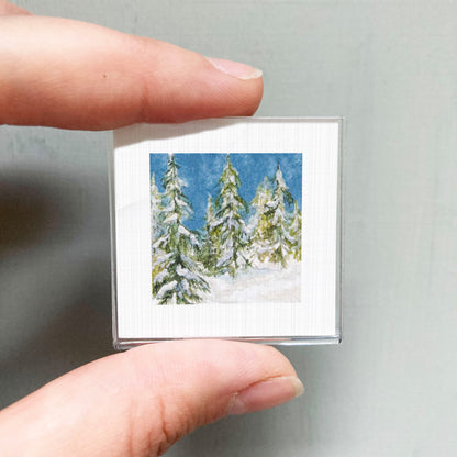 A person's hand holding a small square picture of a snowy winter landscape with pine trees.