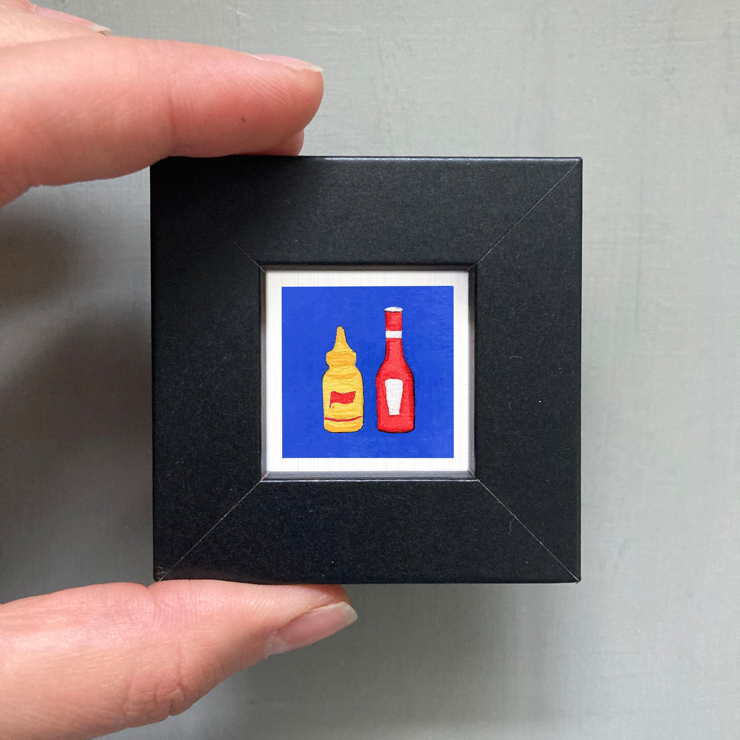 A person's hand is holding a small black square frame containing a framed image of two condiment bottles, one yellow and one red.