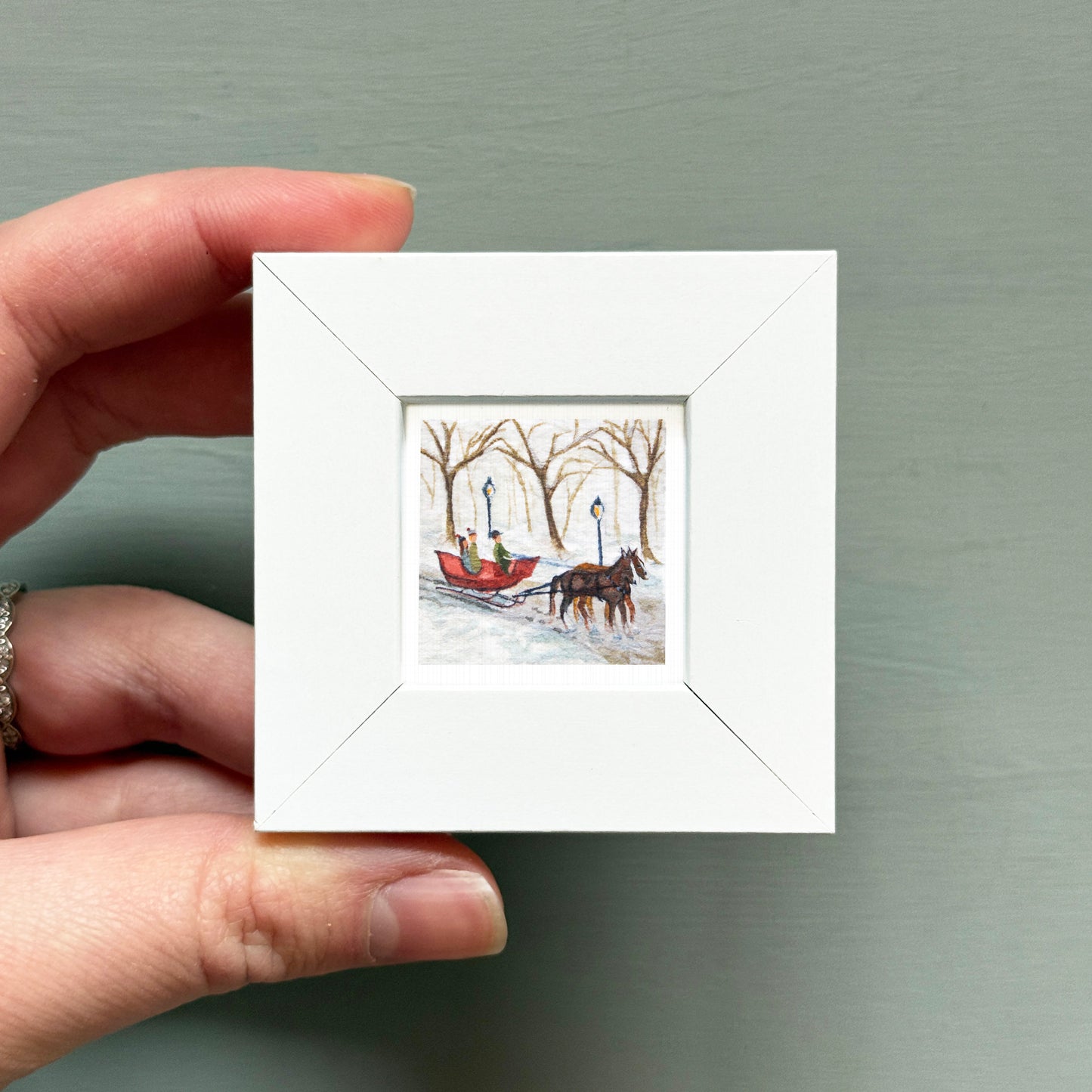 A hand is holding a small white picture frame containing a painting of a horse-drawn sleigh in a snowy forest.