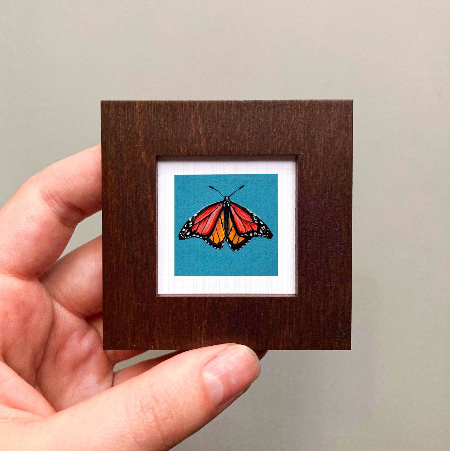 A hand is holding a small wooden frame containing a vibrant, colorful butterfly image against a blue background.