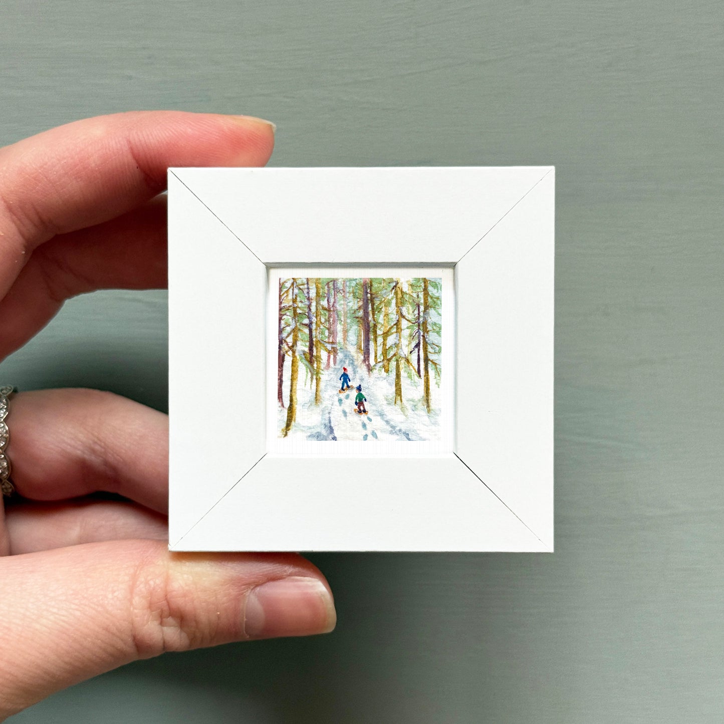 A hand is holding a small white square frame containing a painting of two people skiing in a snowy forest.