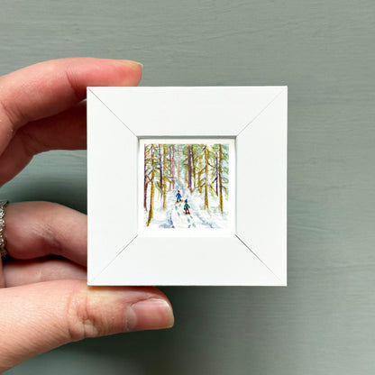 A hand is holding a small white square frame containing a painting of two people skiing in a snowy forest.
