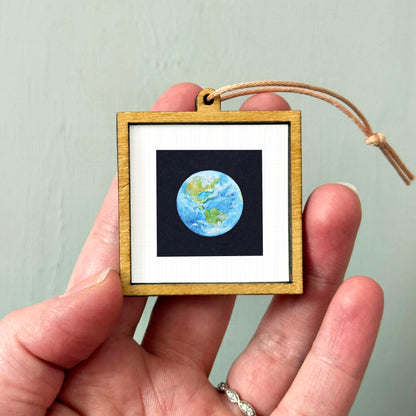 A hand holding a small wooden frame containing a miniature representation of the Earth.