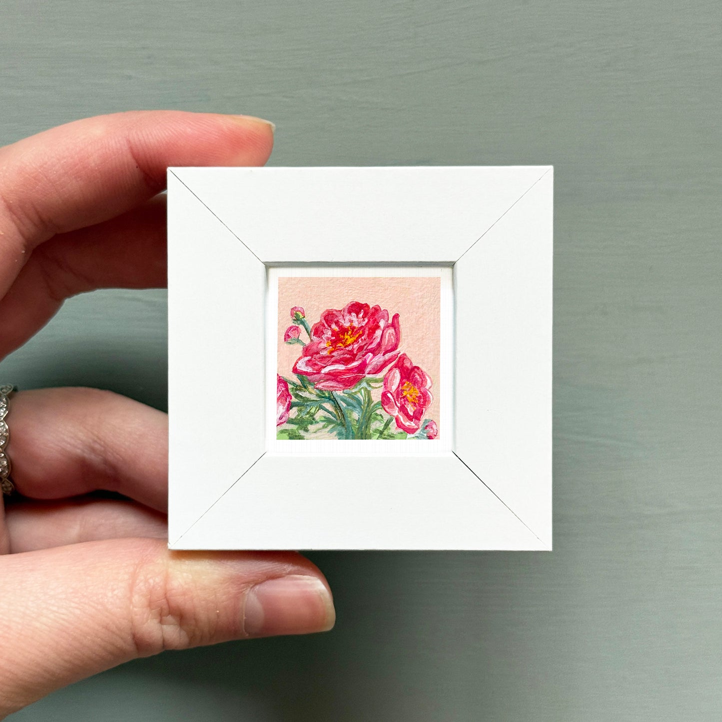A hand is holding a small white square frame containing a vibrant painting of pink flowers.