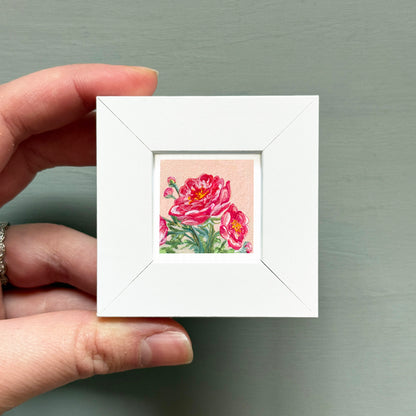 A hand is holding a small white square frame containing a vibrant painting of pink flowers.