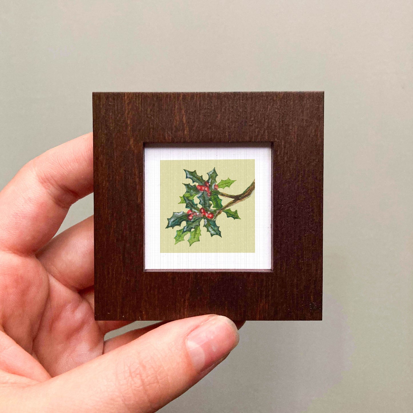 A hand is holding a small wooden frame containing a framed picture of a holly branch with berries.