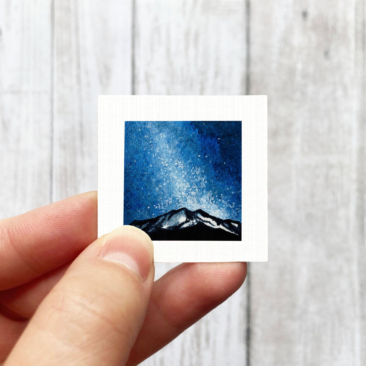 A hand is holding a small square artwork depicting a night sky with stars and a mountain range.