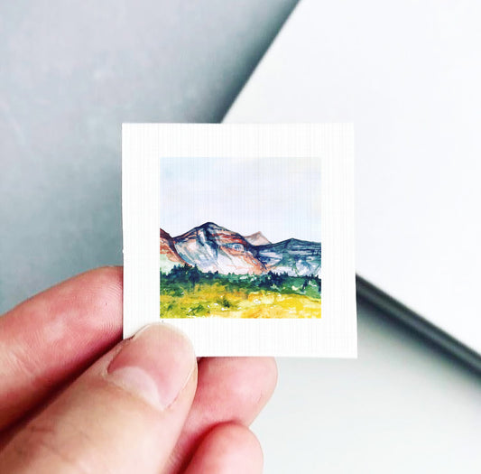 A hand is holding a small square-shaped piece of paper with a watercolor painting of a mountain landscape on it.