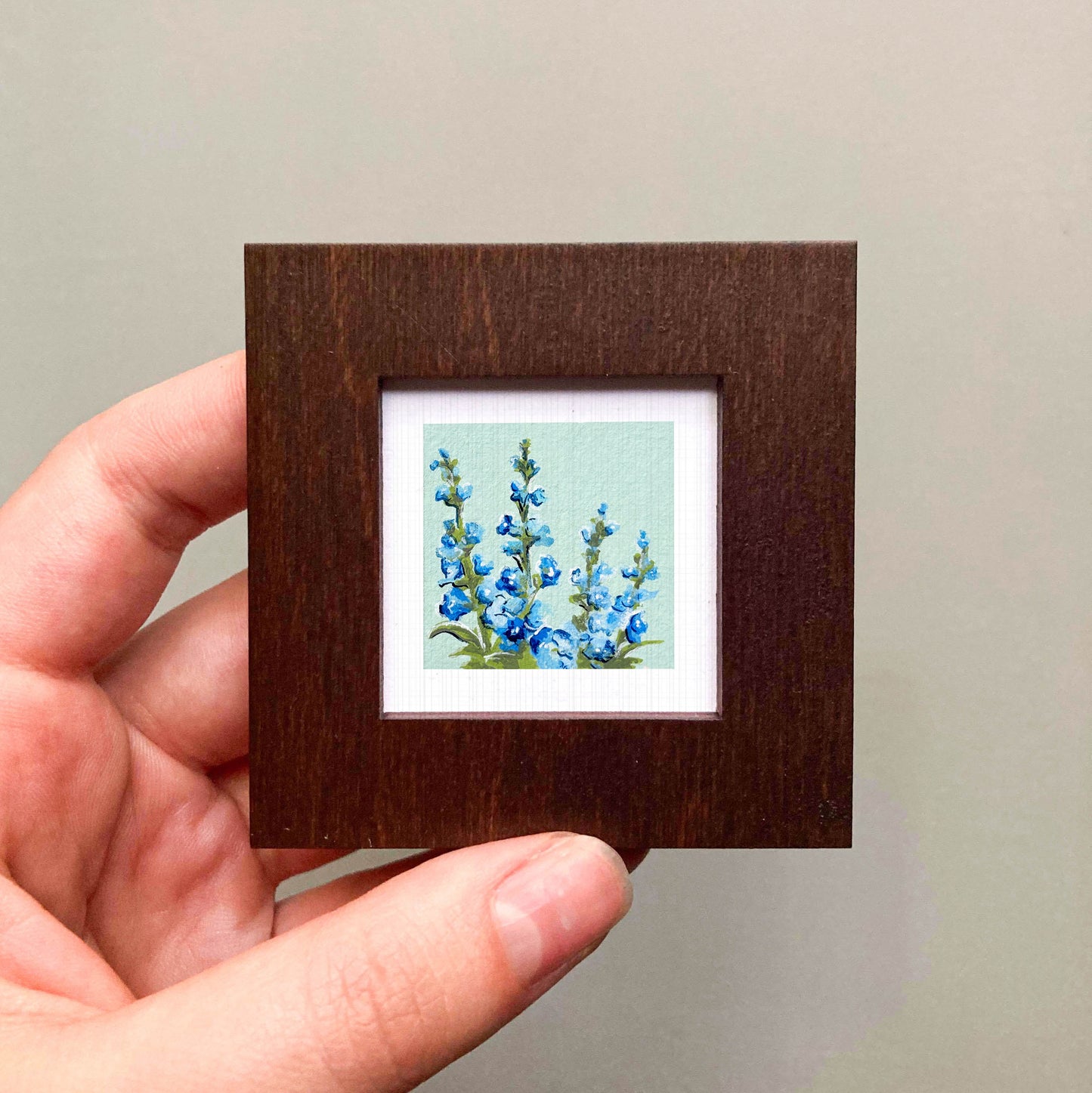 A hand is holding a small wooden frame containing a painting of blue flowers.