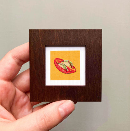 A hand is holding a small wooden frame containing a framed illustration of a red and yellow object, which appears to be a piece of artwork or a decorative item.