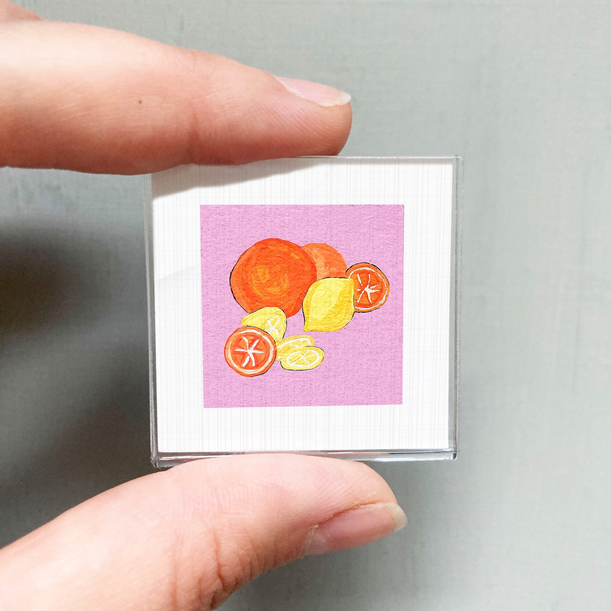 A hand holding a small square with an illustration of various fruits, including oranges and lemons, against a pink background.