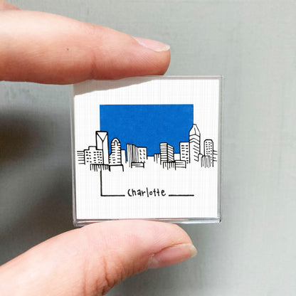A hand holding a small square with a drawing of a city skyline and the word "Charlotte" written below it.