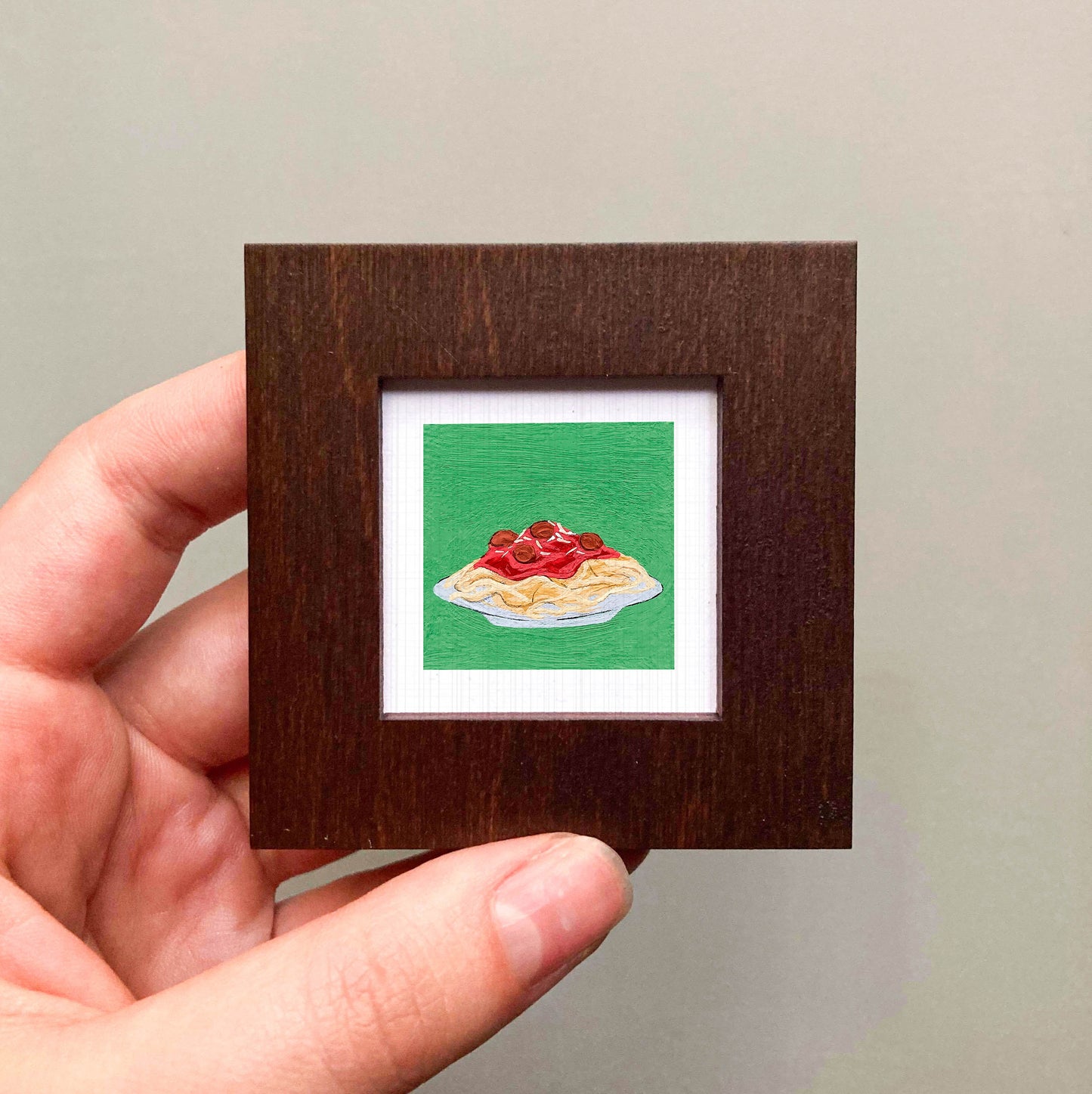 A hand is holding a small wooden frame containing a framed illustration of a plate of spaghetti with red sauce.