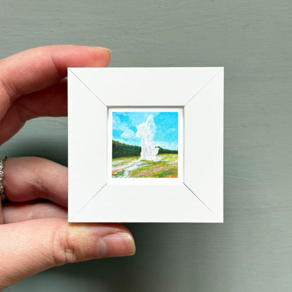 A hand is holding a small white square frame containing a painting of a landscape with a tall structure in the background.