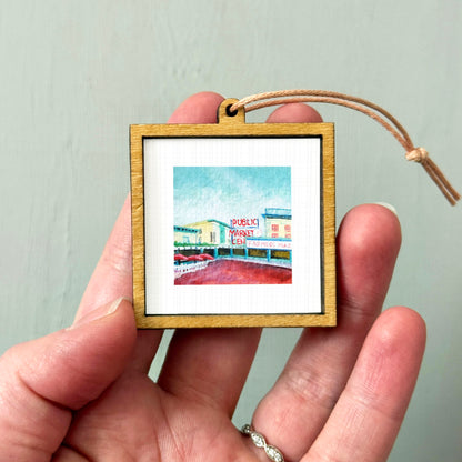 A hand holding a small wooden frame containing a painting of a market scene.