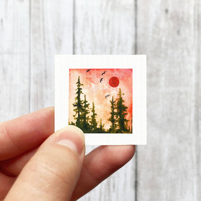 A hand is holding a small square-shaped artwork depicting a forest scene with trees and a red sun in the sky.