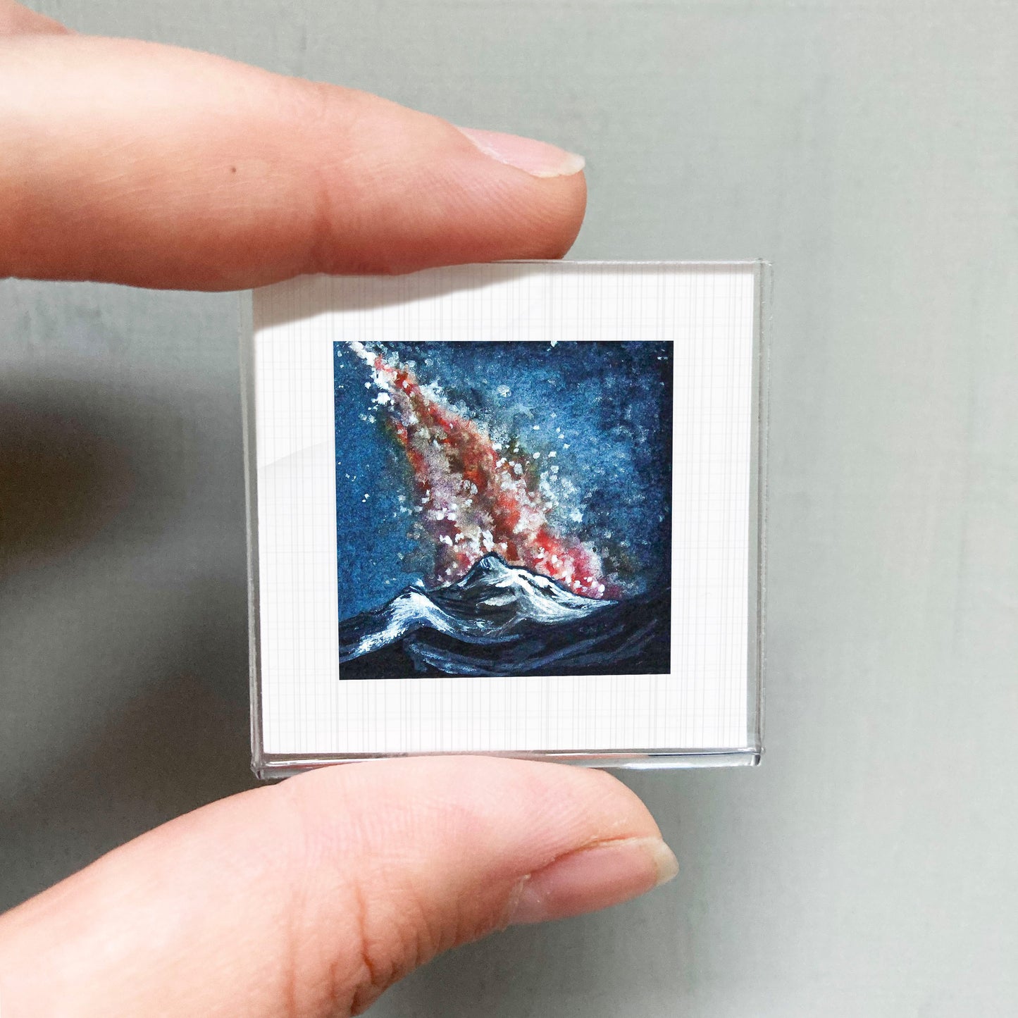 A person's hand is holding a small square picture of a colorful, abstract painting depicting a wave crashing against a mountain.