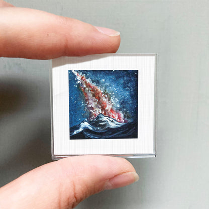 A person's hand is holding a small square picture of a colorful, abstract painting depicting a wave crashing against a mountain.