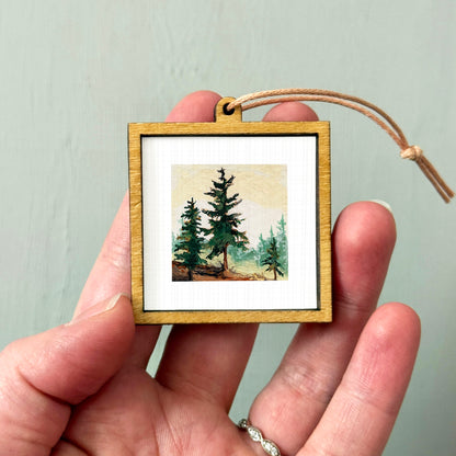 A hand holding a small wooden frame containing a painting of pine trees.