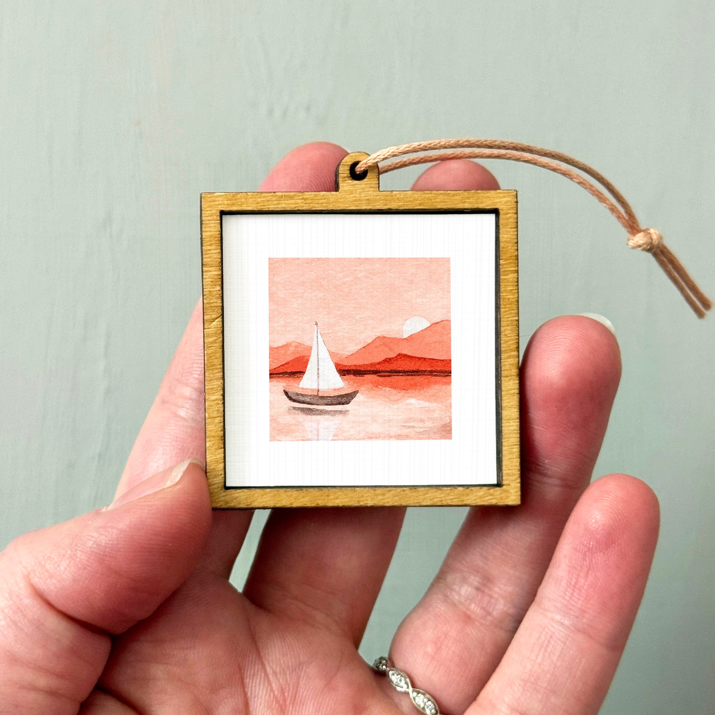 A hand holding a small wooden frame containing a picture of a sailboat on a body of water.