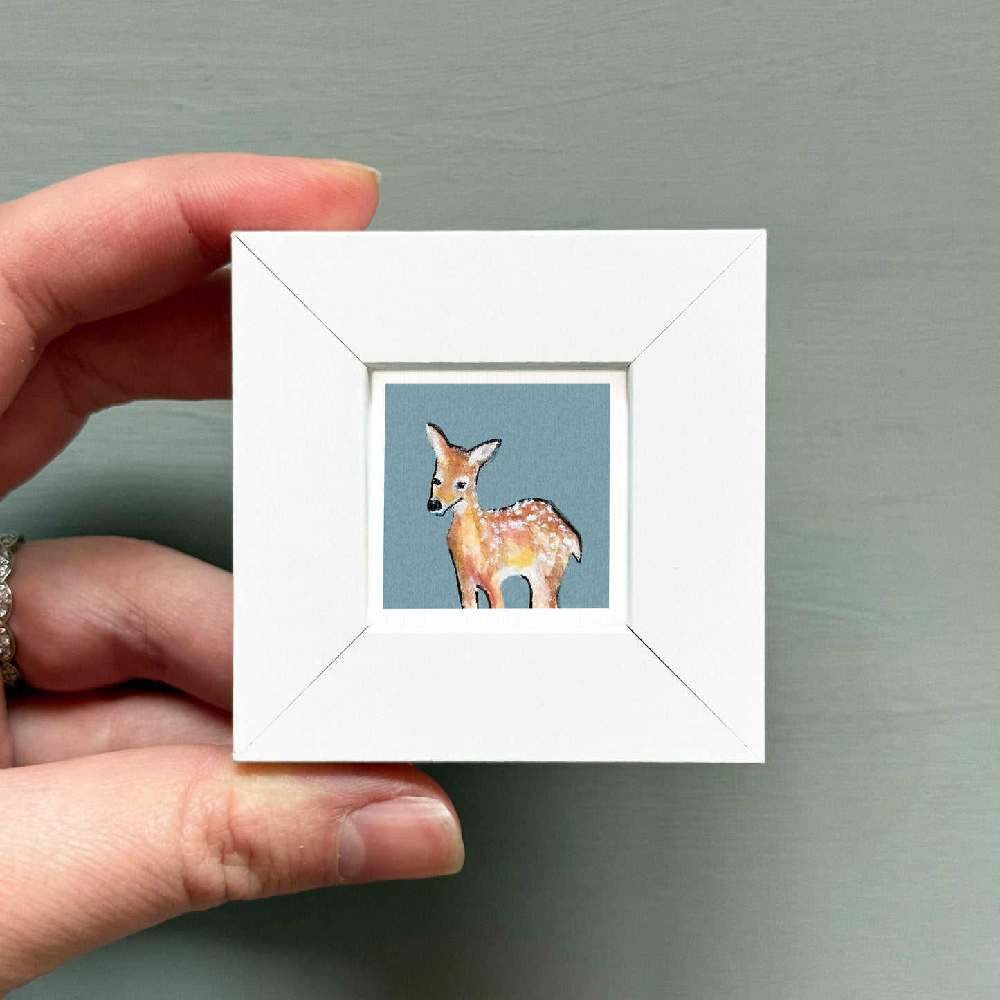 A hand is holding a small white picture frame containing a painting of a deer.
