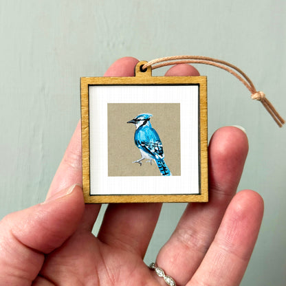 A hand holding a small wooden frame containing a picture of a blue jay.