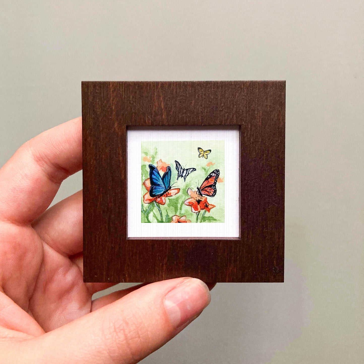 A hand is holding a small, square-shaped picture frame containing a painting of colorful butterflies on flowers.