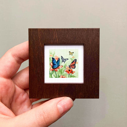 A hand is holding a small, square-shaped picture frame containing a painting of colorful butterflies on flowers.