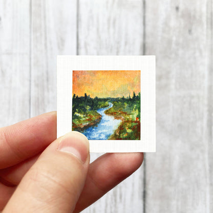 A hand is holding a small square painting of a river with trees and a sky background.