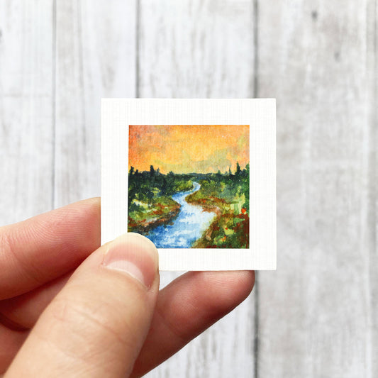 A hand is holding a small square painting of a river with trees and a sky background.