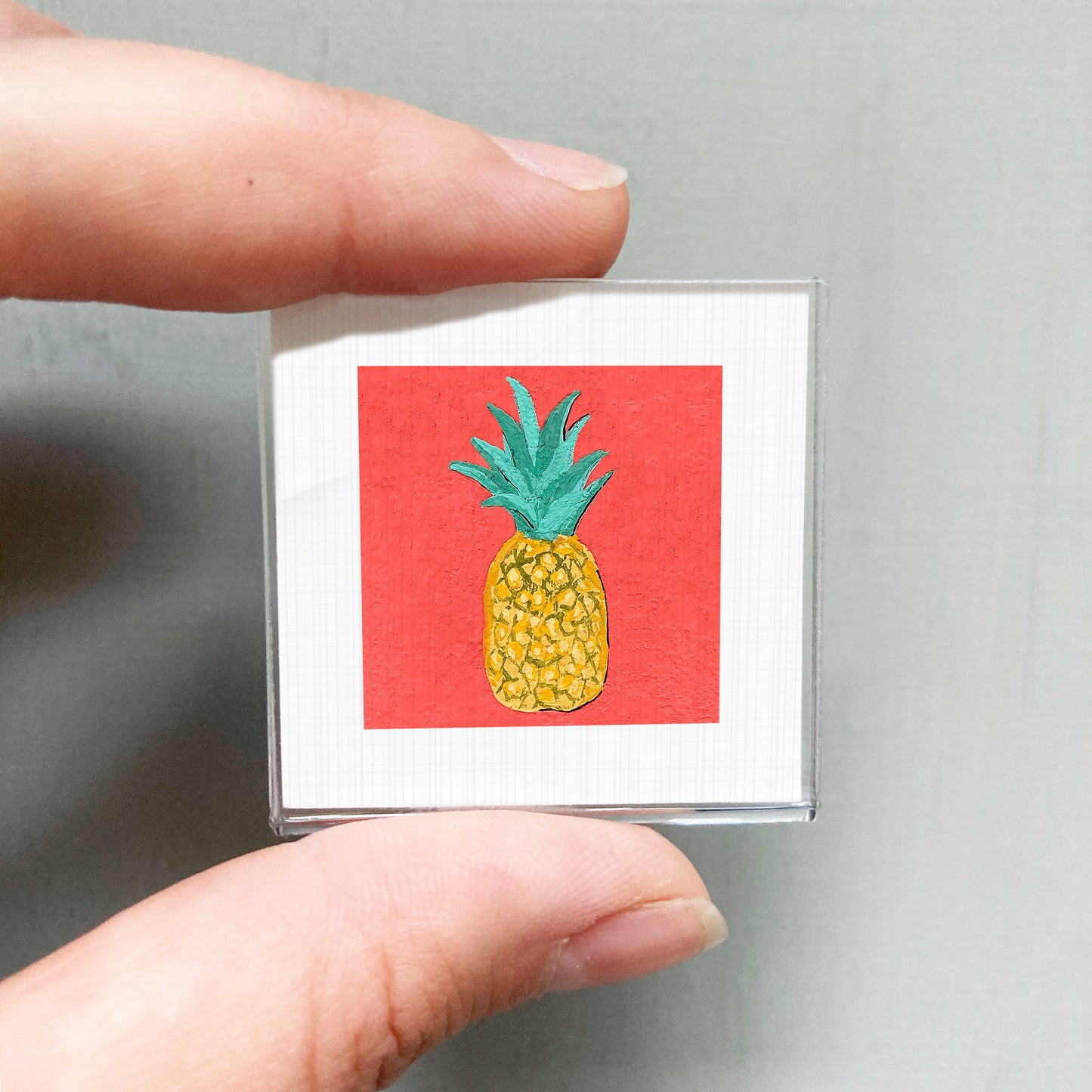 A person's hand holding a small square with a vibrant red background and a pineapple illustration.