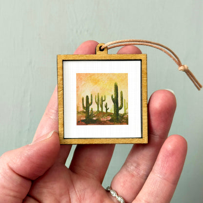 A hand holding a small wooden frame containing a painting of cacti.