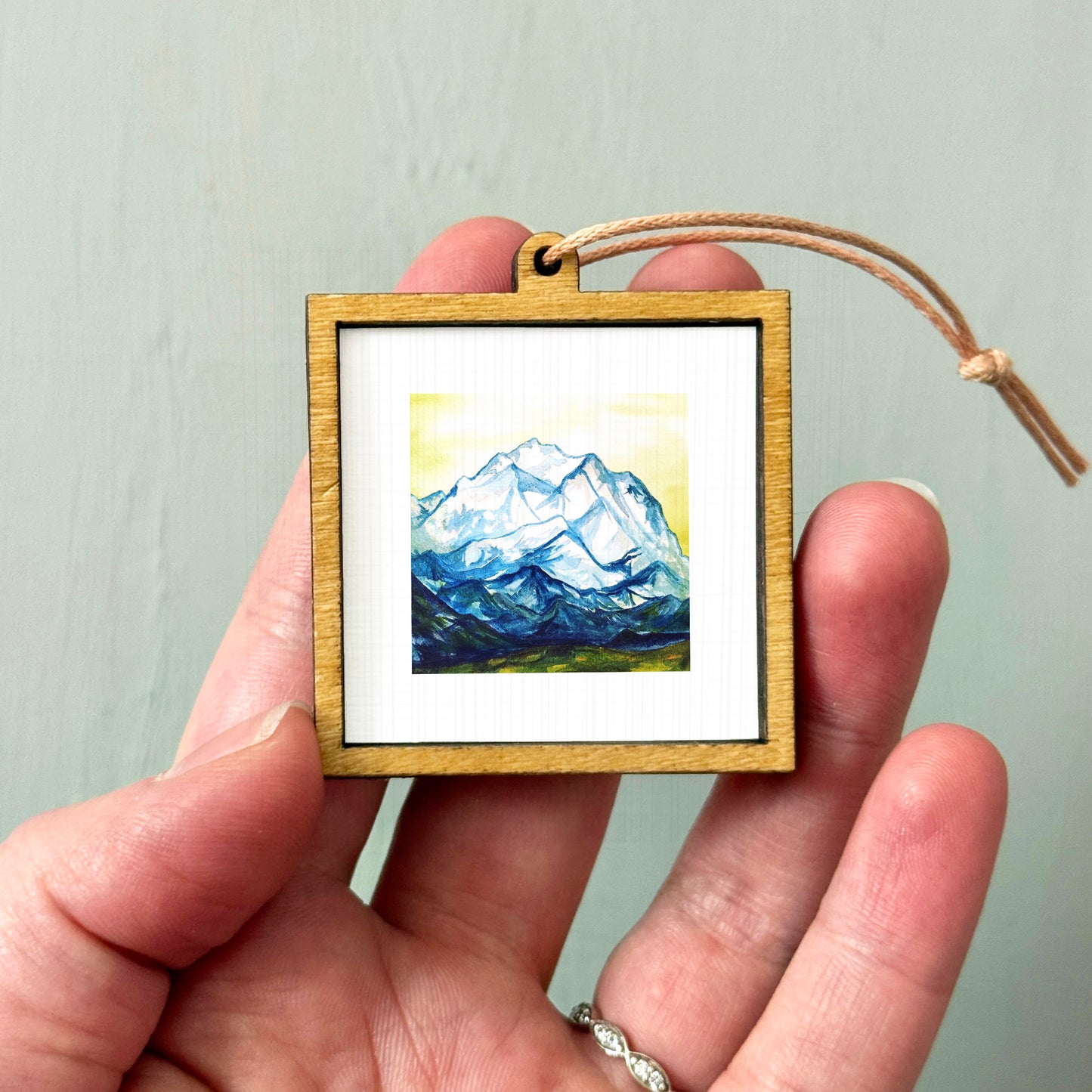 A person's hand holding a small wooden frame containing a framed painting of a mountain landscape.